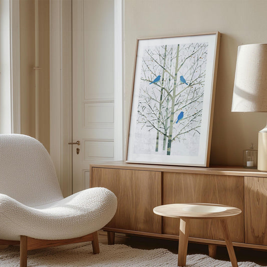 Framed art print of three bluebirds perched on bare winter branches, displayed on a light wood credenza in a cozy modern living room. The space features a cream textured lounge chair, matching rug, small round wood table, and soft neutral tones, creating a serene Scandinavian-inspired aesthetic. Natural light filters through a tall window beside the artwork.