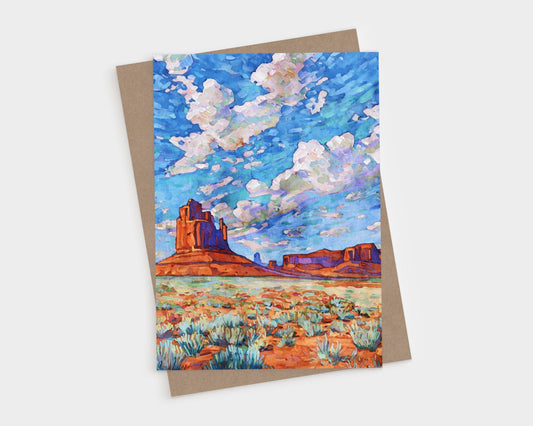 Vibrant greeting card featuring a painting of Monument Valley. The scene shows towering red rock buttes rising from a desert landscape dotted with green sagebrush. Above, a dramatic sky of vivid blues and purples is filled with fluffy white clouds, painted in bold, impressionistic strokes. The artwork captures the vast, colorful beauty of the American Southwest.