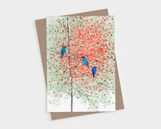 Watercolor greeting card featuring three bright bluebirds perched on thin branches of a maple tree with leaves transitioning from green to autumn red. The soft, impressionistic background creates a serene seasonal atmosphere. Card shown with a brown kraft envelope on a white background.
