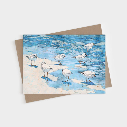 Watercolor-style illustration of small white shorebirds with orange legs standing and wading at the edge of blue ocean waves on a sandy beach, printed on a folded greeting card with a kraft brown envelope behind it.