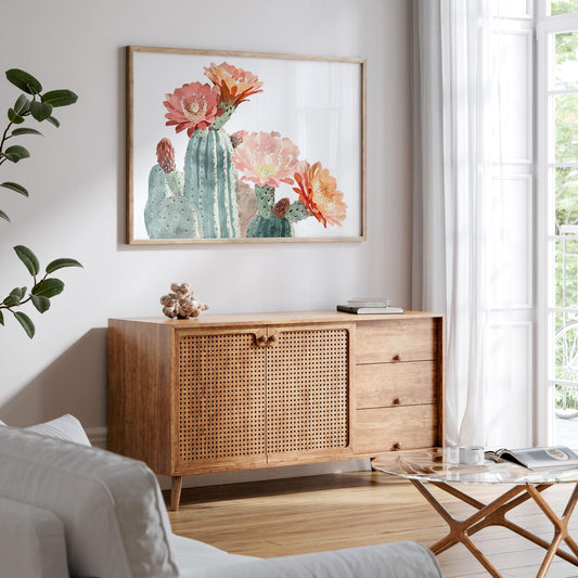 Framed watercolor cactus print with coral, peach, and orange flowers hangs above a wooden sideboard in a bright living room with natural light, white curtains, and minimalist decor.