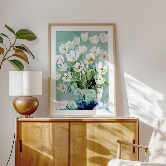 Framed art print of white tulips in a blue-green vase hangs above a light wood sideboard in a sunlit modern living room. The floral painting features soft teal and green hues with layered watercolor textures. A mid-century style lamp and leafy plant complete the warm, minimalist decor.