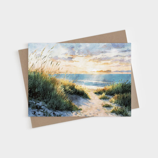 Watercolor greeting card featuring a sandy path through tall beach grass leading to the ocean at sunset, with warm golden light reflecting on the waves and clouds. A kraft envelope is placed behind the card.