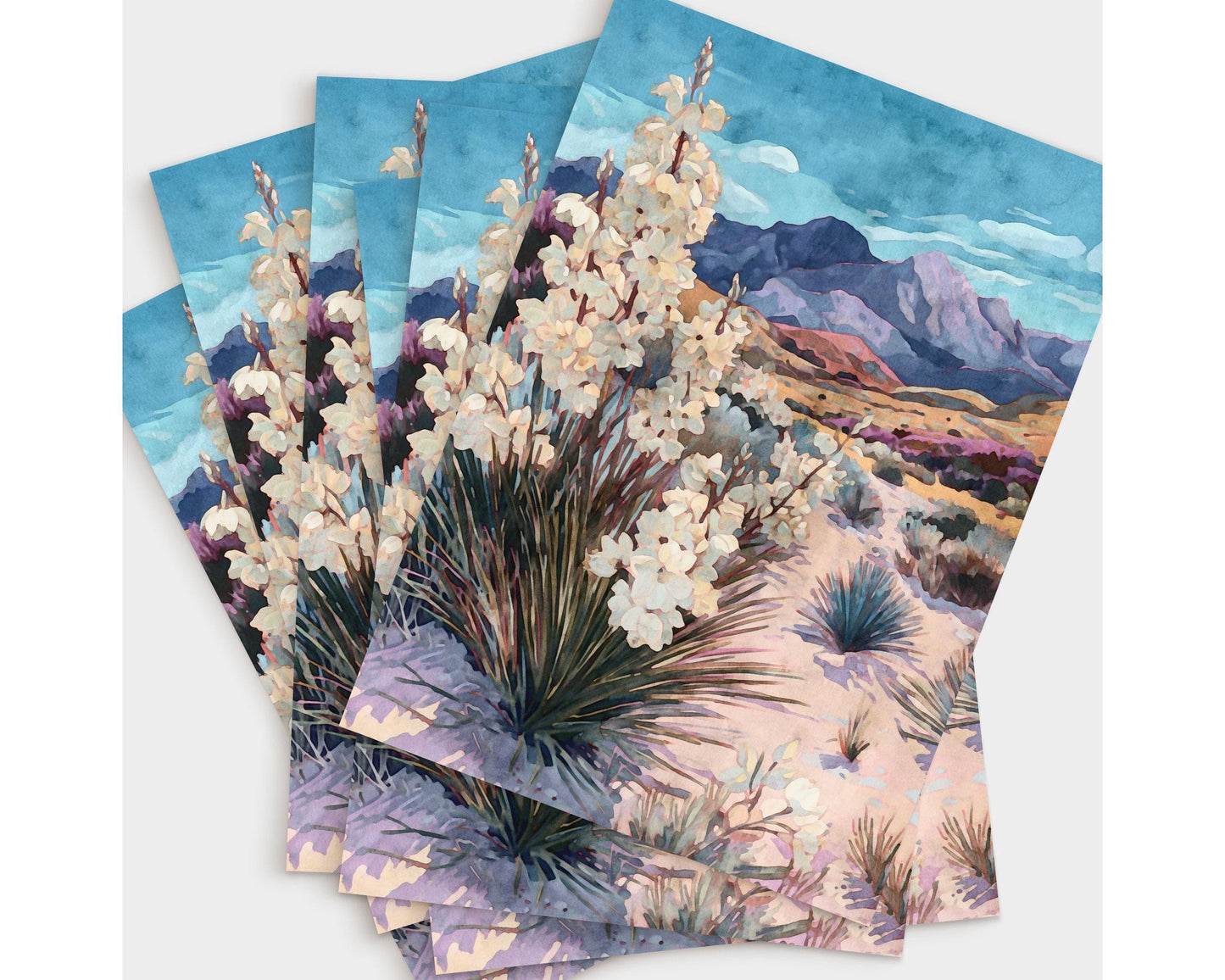 Set of greeting cards arranged in a fanned stack, each featuring a watercolor desert landscape with blooming yucca plants in the foreground, sandy terrain, and purple mountains under a bright blue sky with soft clouds.