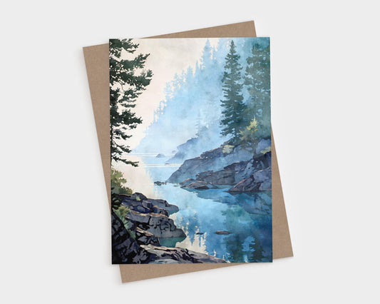 Vertical art greeting card featuring a watercolor-style misty lakeshore scene with calm blue water reflecting evergreen trees and rocky shoreline. Soft morning fog drifts through layered pine trees along a quiet inlet, painted in cool blue and green tones. Shown with a natural kraft paper envelope on a clean white background.