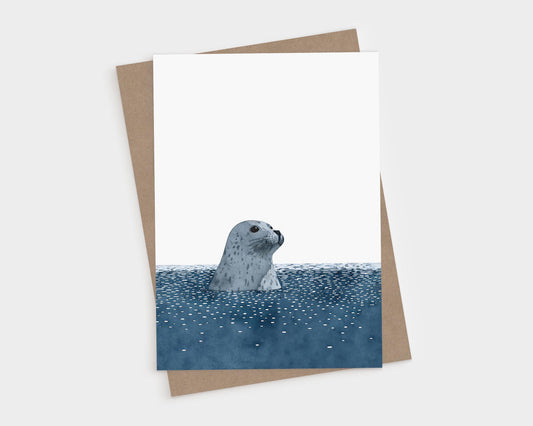 Minimalist greeting card featuring a watercolor-style illustration of a gray spotted seal with its head poking above deep blue water, set against a clean white background. A brown kraft envelope is placed behind the card.