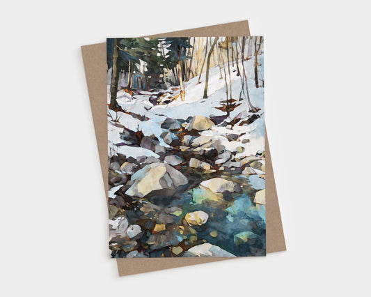 Watercolor-style greeting card featuring a rocky creek running through a snowy forest. Sunlight filters through bare trees, reflecting on the icy water in shades of blue and green. The card is displayed with a kraft paper envelope.