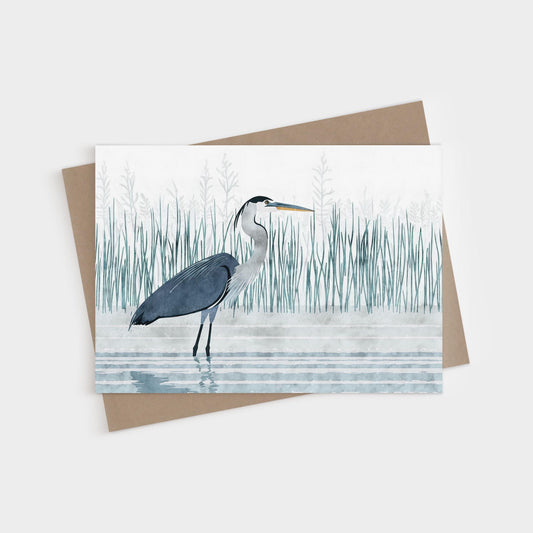 Greeting card featuring a minimalist illustration of a great blue heron standing in shallow water, surrounded by tall, delicate marsh grasses in muted blue-green tones. A kraft brown envelope is partially visible beneath the card.