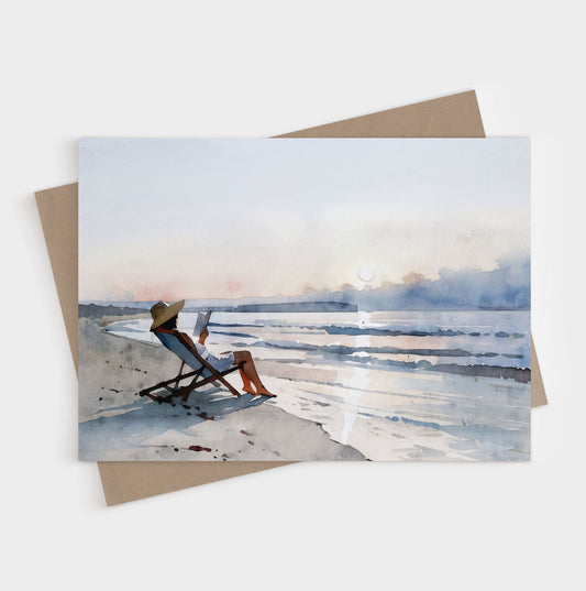 Greeting card and kraft envelope with a watercolor-style illustration of a woman lounging in a beach chair reading a book at sunset, with soft waves and pastel sky in the background.