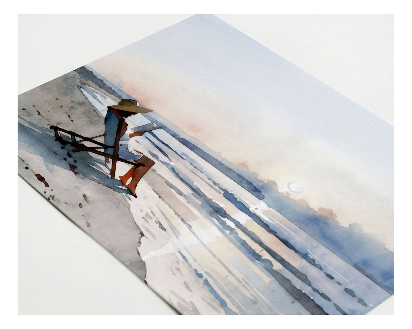 Angled close up of a greeting card with a watercolor-style illustration of a woman lounging in a beach chair reading a book at sunset, with soft waves and pastel sky in the background.