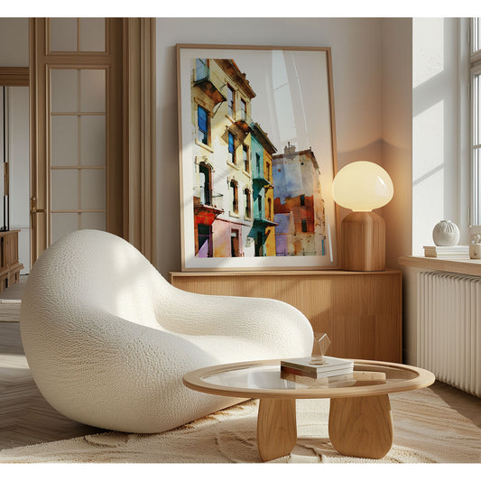 Framed watercolor painting of colorful buildings with pastel facades, leaning against a wall in a sunlit room with a modern chair and wooden table.