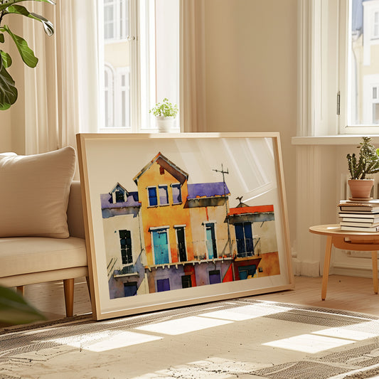 Framed watercolor painting of colorful buildings leaning against a beige sofa in a sunlit living room with plants and books.