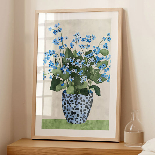 Framed artwork featuring a watercolor-style depiction of a bouquet of blue forget-me-not flowers with green leaves, arranged in a patterned blue vase, leaning against a wall on a wooden surface in a softly lit room.
