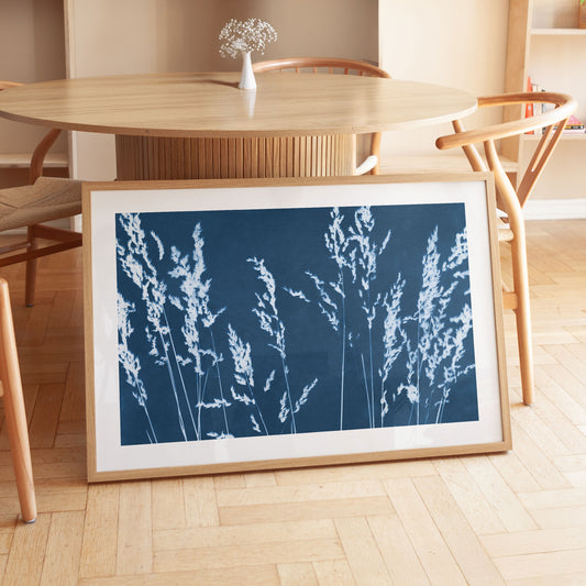Framed cyanotype print of wild grasses leaning against a wooden dining table, featuring white botanical silhouettes on a deep blue background.