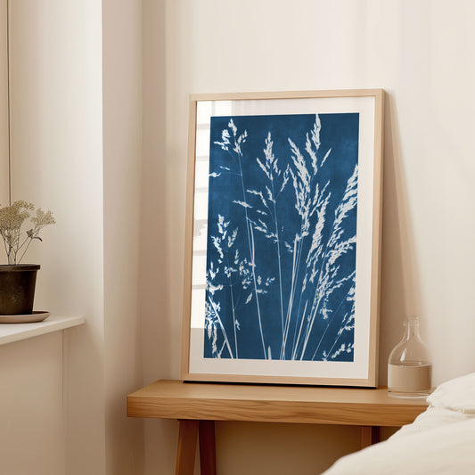 Framed cyanotype print of wild grasses leaning against a wall on a wooden side table, featuring white botanical silhouettes on a deep blue background.