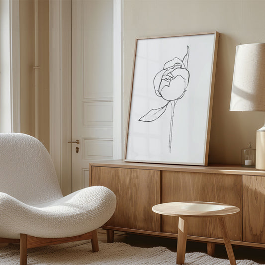 Framed black and white line drawing of a blooming peony flower, leaning against a beige wall on a wooden sideboard in a modern interior.
