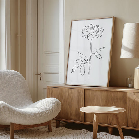 Framed minimalist line drawing of a peony flower leaning against a wall on a wooden sideboard, with a modern beige chair and soft lighting in the room.