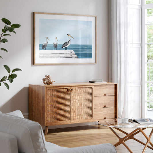 Framed watercolor-style artwork of three pelicans on a wooden dock over a calm ocean, displayed above a mid-century modern wood cabinet in a bright, coastal-inspired living room.