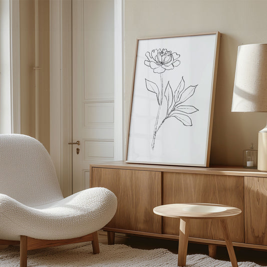 Framed minimalist line drawing of a blooming peony with detailed petals and leaves, leaning against a beige wall on a wooden sideboard.