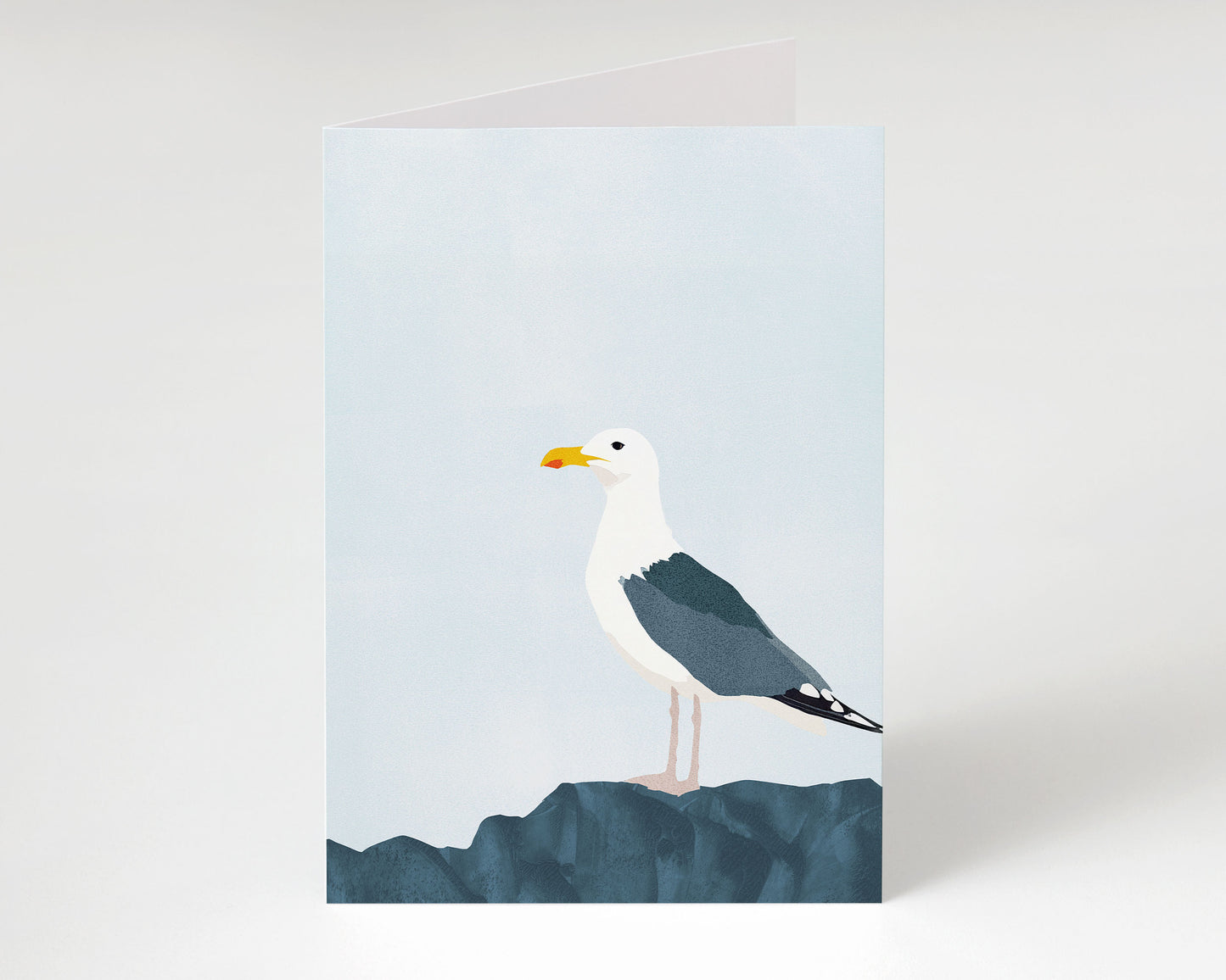 Minimalist greeting card standing upright, featuring a seagull with dark wings and white head perched on textured blue rocks beneath a pale sky.