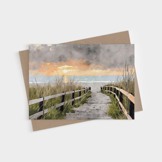Greeting card featuring a watercolor-style painting of a wooden boardwalk path leading through grassy dunes to the ocean, with a glowing orange and yellow sunset under dramatic gray clouds.