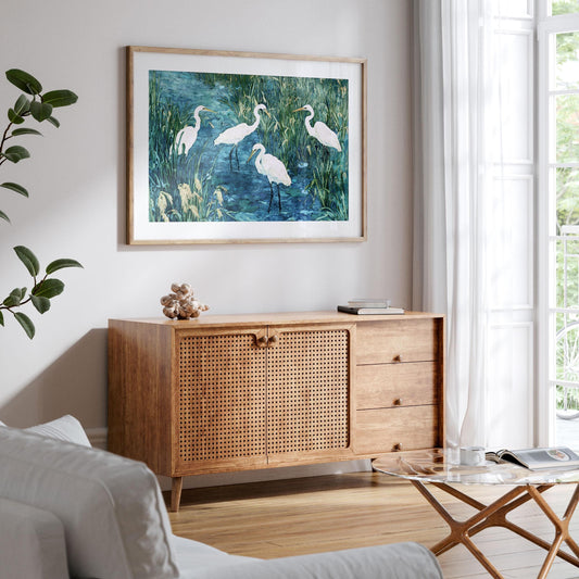 Framed painting of five white egrets in a marsh, with blue water and tall green grasses, hangs above a mid-century style wood sideboard with woven rattan doors. The bright, airy living room features a white sofa, glass coffee table, books, and large windows with sheer curtains, creating a calm, natural atmosphere.