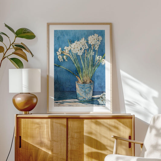 Framed watercolor-style art print of white narcissus flowers in a blue ceramic pot, set against a deep blue background. The artwork hangs above a wooden sideboard, styled with a modern brown lamp and a leafy plant. Natural sunlight casts soft shadows across the wall and furniture, creating a warm, minimalist interior scene.
