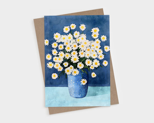 Greeting card featuring a watercolor illustration of white daisies with yellow centers in a blue polka dot vase, set against a dark blue background and light blue tabletop. A kraft brown envelope is placed beneath the card.