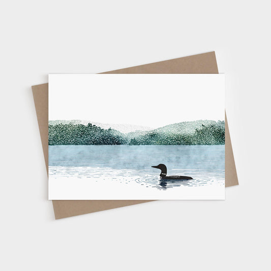 Illustrated greeting card featuring a loon gliding across a calm lake with rippling water. Behind the bird, layers of misty green forested hills fade into the distance under a wide white sky. The artwork is minimal and serene, with soft watercolor textures. Card is paired with a natural kraft envelope.