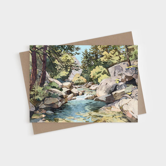 Greeting card featuring a watercolor-style painting of a mountain stream flowing between large boulders and pine trees, with sunlight filtering through green foliage. The card is displayed with a brown kraft envelope on a white background.