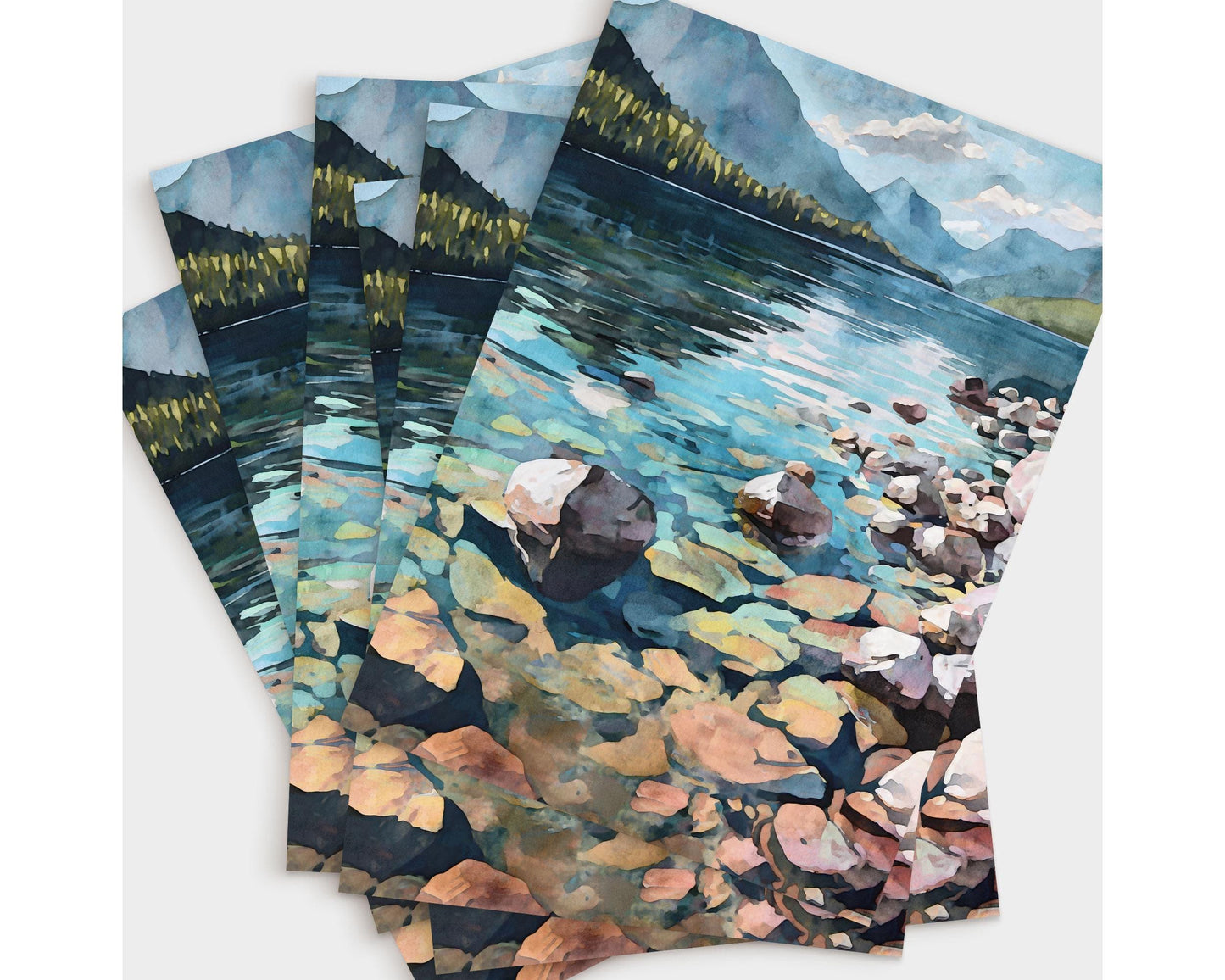 Set of several greeting cards featuring a watercolor-style illustration of a mountain lake with clear water, colorful rocks, a rocky shoreline, forested hills, and distant mountains under a partly cloudy sky, fanned out to show the repeated scenic design.