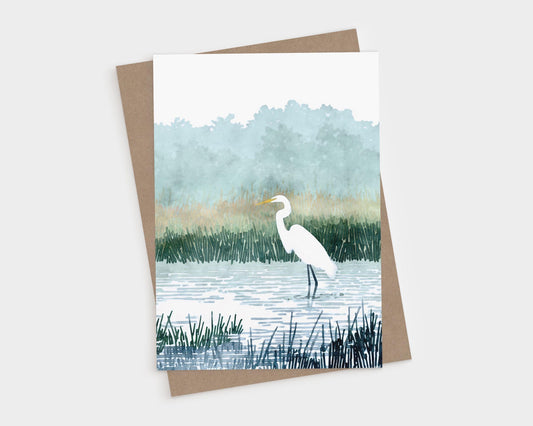 Watercolor-style greeting card featuring a white egret standing in calm marsh waters surrounded by tall grasses, with soft green trees and mist in the background.