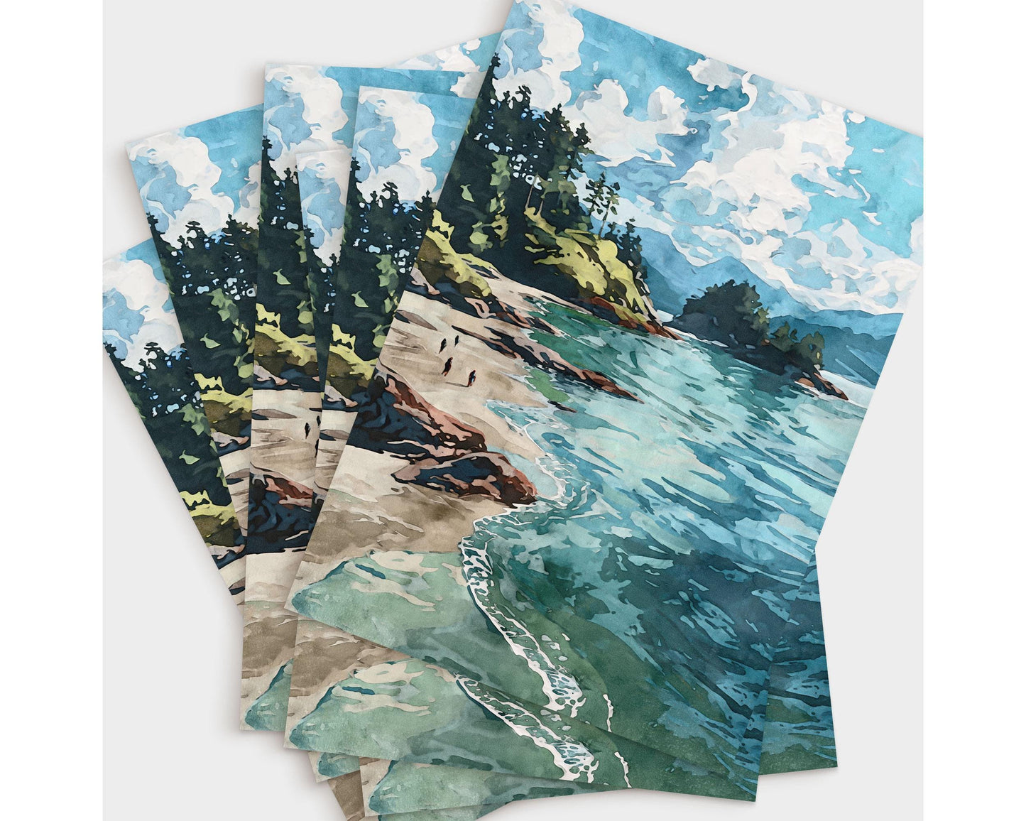 Set of greeting cards featuring a watercolor-style coastal scene with turquoise waves, sandy beach, rocky shoreline, and pine-covered cliffs beneath a bright blue sky with fluffy white clouds, fanned out in a layered display.