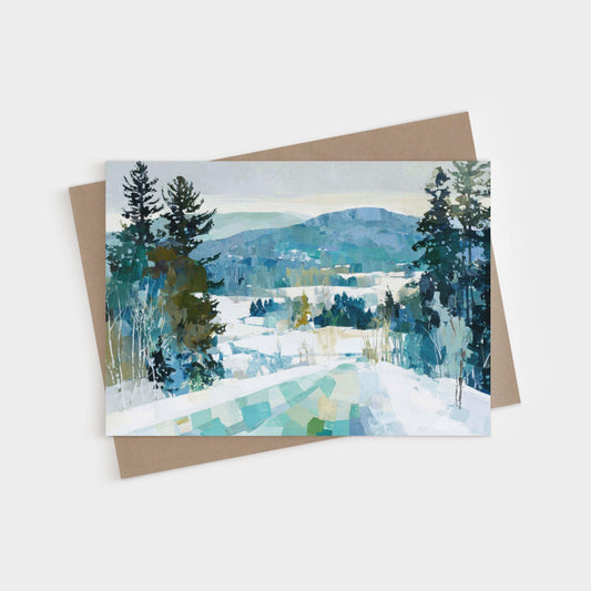 Greeting card featuring a winter landscape painting of snow-covered hills, pine trees, and a distant mountain range in cool blue, green, and white tones. Card is displayed with a kraft brown envelope.