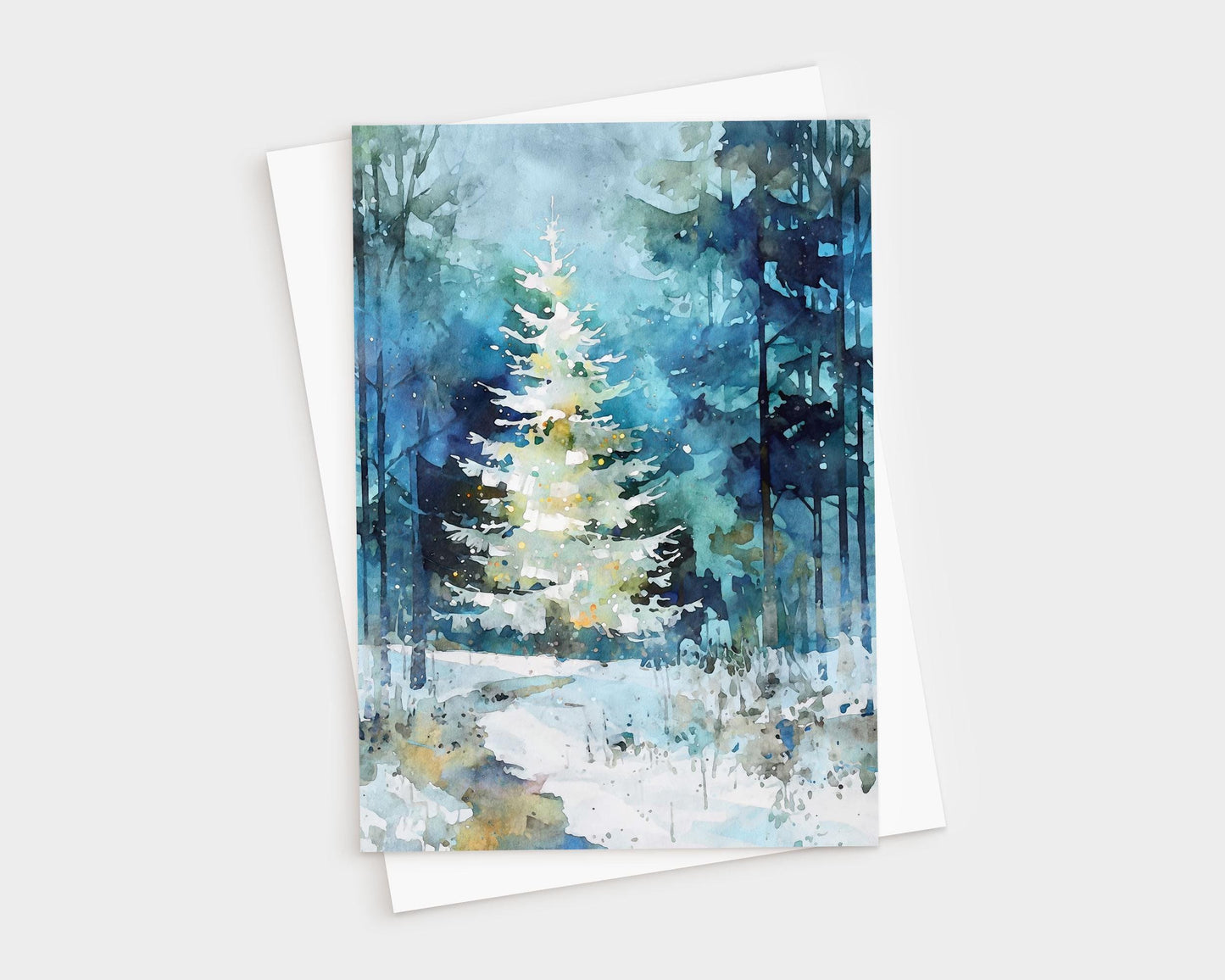 A folded greeting card featuring a watercolor-style painting of a glowing white Christmas tree in a snowy blue-green forest, shown with a white envelope. Soft washes, tall tree silhouettes, and gentle light speckles create a serene, wintry holiday scene.