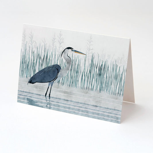 Folded greeting card with an illustration of a great blue heron wading in rippling water, surrounded by tall marsh grasses in soft blue tones. The card stands against a white background, angled to show its clean fold and blank interior.