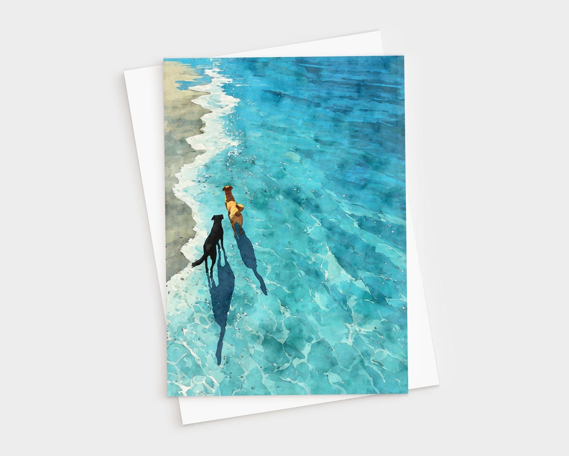 Watercolor-style greeting card featuring two dogs walking along the shoreline, their shadows stretching across the shallow turquoise waves. The sandy beach contrasts with the vivid blue water, creating a peaceful, sunlit scene. A white envelope is partially visible behind the card.