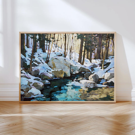 Framed painting of a winter forest scene, showing tall trees, large rocks, and a stream reflecting icy blues and warm golden light. Snow blankets the ground and boulders, while sunlight filters through the trees, creating a serene, peaceful atmosphere. The artwork leans against a white wall on a wooden herringbone floor.