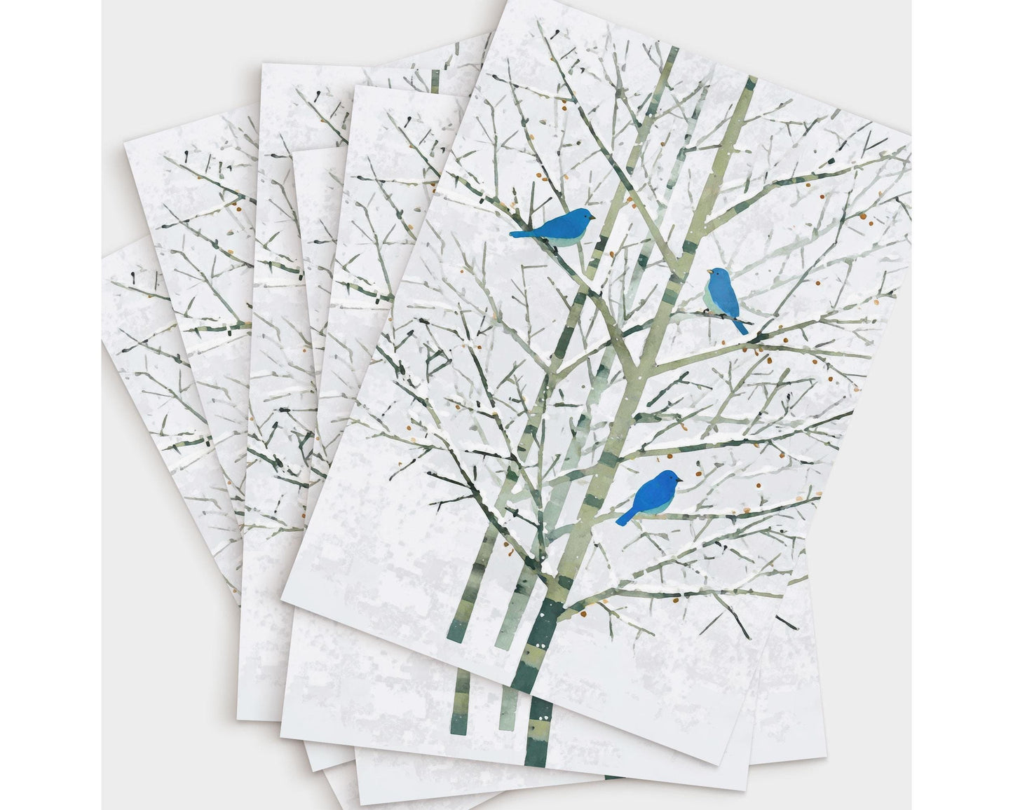 Set of greeting cards featuring a minimalist watercolor-style illustration of three bluebirds perched on pale winter branches. The cards are fanned out on a white background, showcasing the delicate green-gray trees and vivid blue birds that create a serene, nature-inspired design.