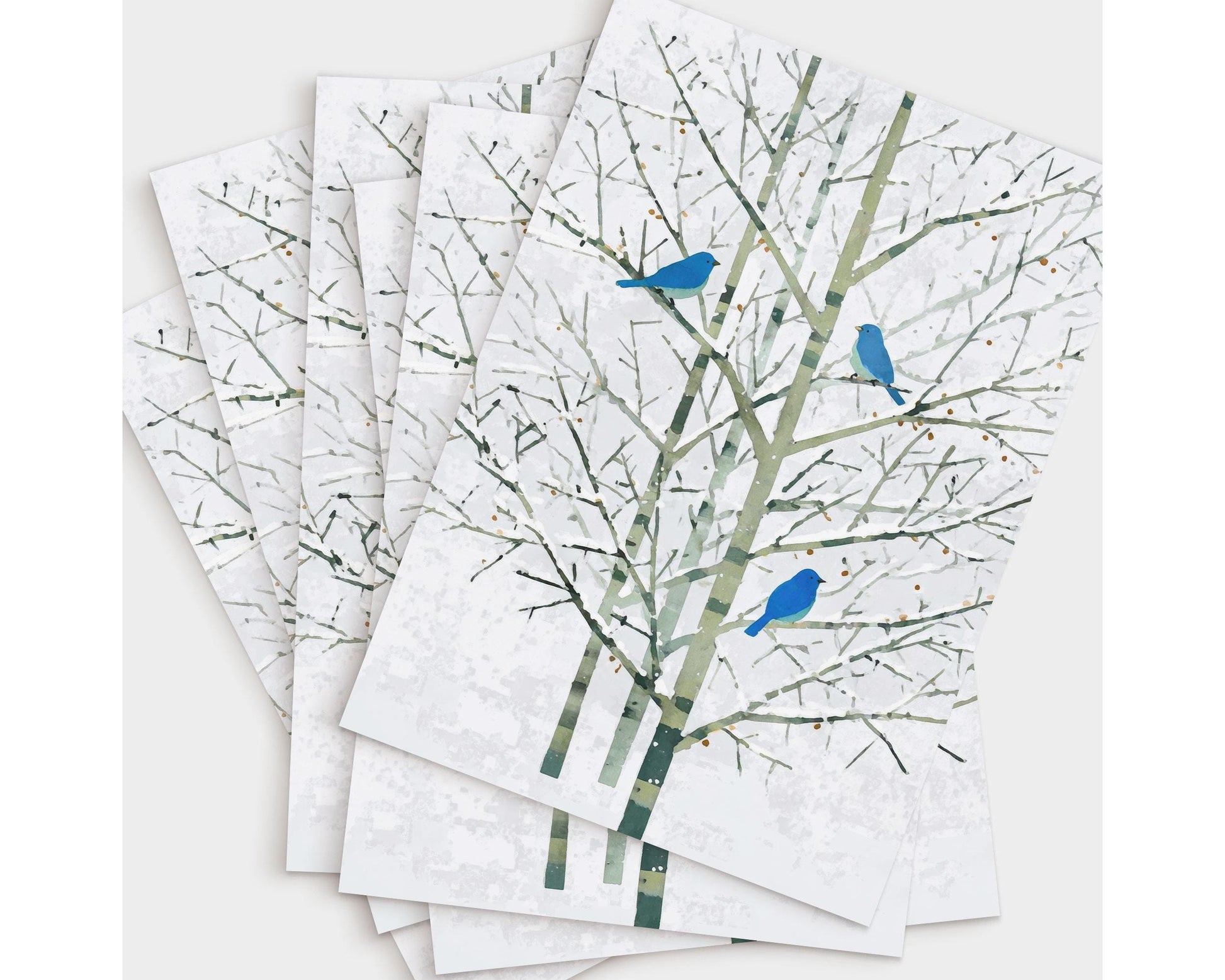 Set of greeting cards featuring a minimalist watercolor-style illustration of three bluebirds perched on pale winter branches. The cards are fanned out on a white background, showcasing the delicate green-gray trees and vivid blue birds that create a serene, nature-inspired design.