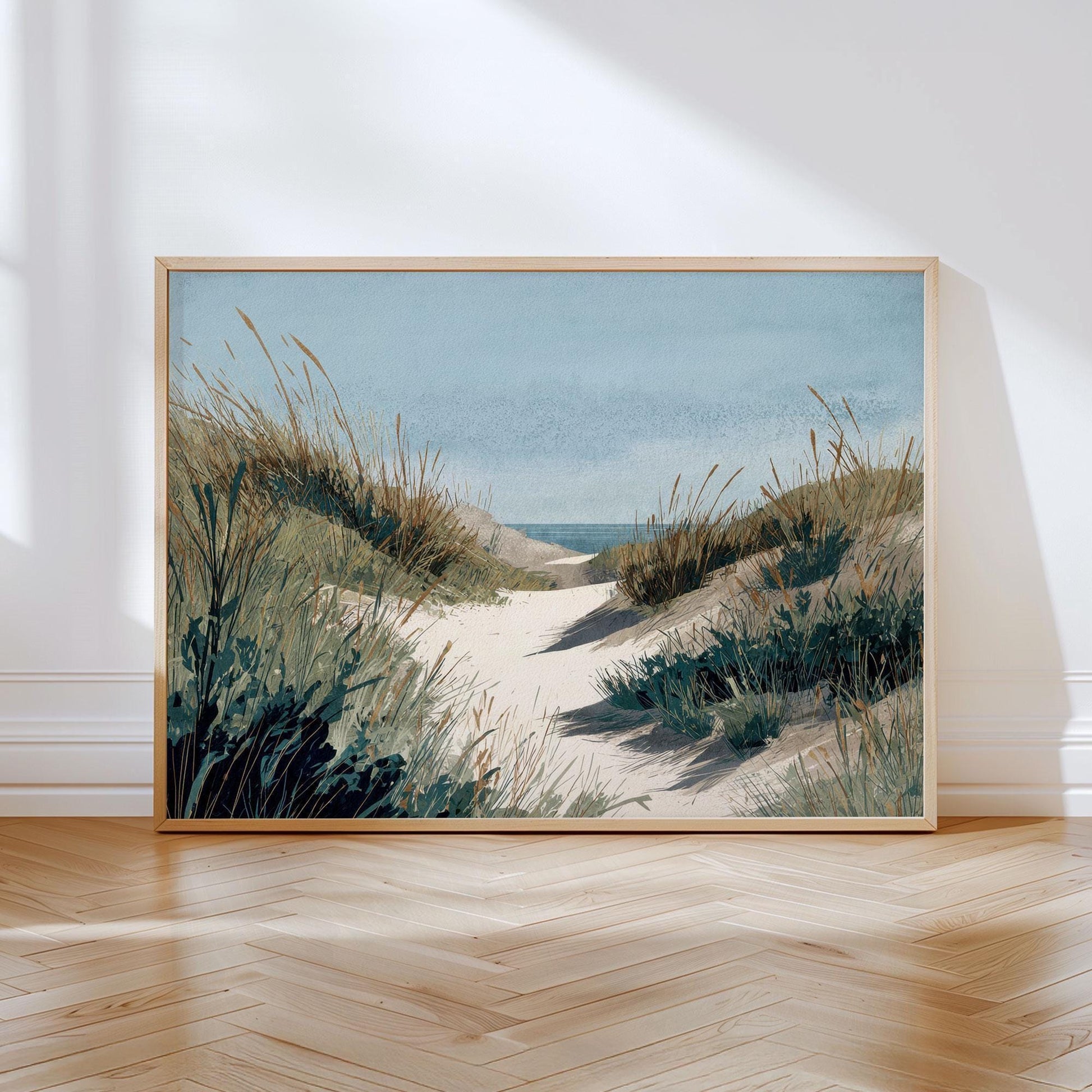 Framed coastal landscape print depicting a sandy path winding through grassy dunes toward the ocean, painted in soft watercolor tones of blue, green, and beige, displayed leaning against a wall on a sunlit wooden floor.