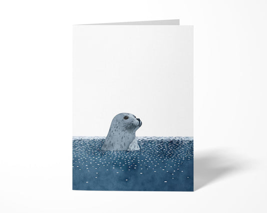 Folded greeting card featuring a minimalist watercolor-style illustration of a gray spotted seal peeking above deep blue, dotted water, set against a clean white background.