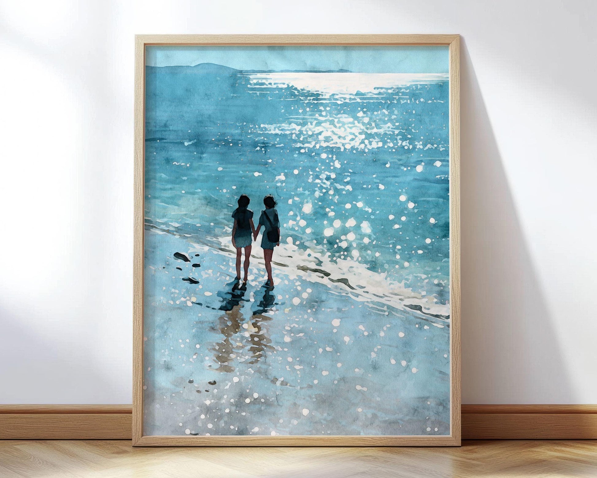 Framed art print featuring a watercolor-style painting of two women walking hand in hand along the shore, with sunlight sparkling on the waves and casting soft reflections on the wet sand. The scene is rendered in cool shades of blue and aqua, evoking a dreamy, peaceful beach atmosphere.