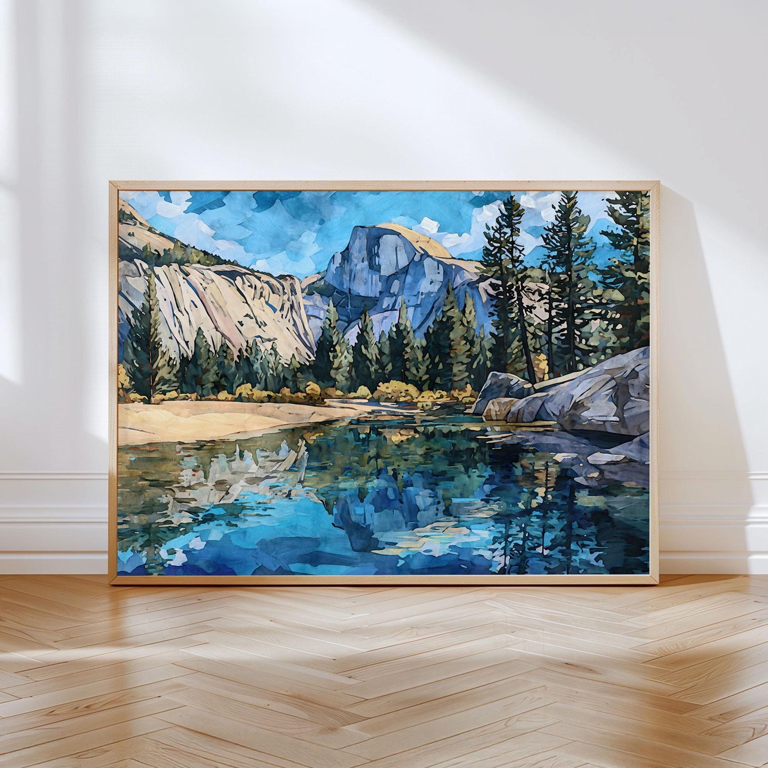 Framed art print of Yosemite’s Half Dome with pine trees and rocky cliffs reflected in a calm blue river, painted in a modern impressionist watercolor style. The artwork rests against a white wall on a light wood herringbone floor, lit by soft natural sunlight.