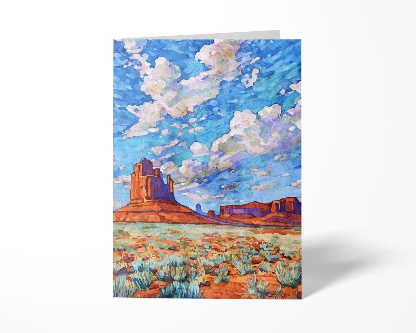 Greeting card featuring a vivid Monument Valley painting. The artwork depicts red rock buttes and mesas against a desert floor dotted with sagebrush, beneath a dramatic sky filled with swirling clouds in shades of blue, lavender, and white. The card stands upright on a white background, highlighting its colorful design.