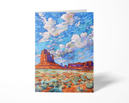 Greeting card featuring a vivid Monument Valley painting. The artwork depicts red rock buttes and mesas against a desert floor dotted with sagebrush, beneath a dramatic sky filled with swirling clouds in shades of blue, lavender, and white. The card stands upright on a white background, highlighting its colorful design.