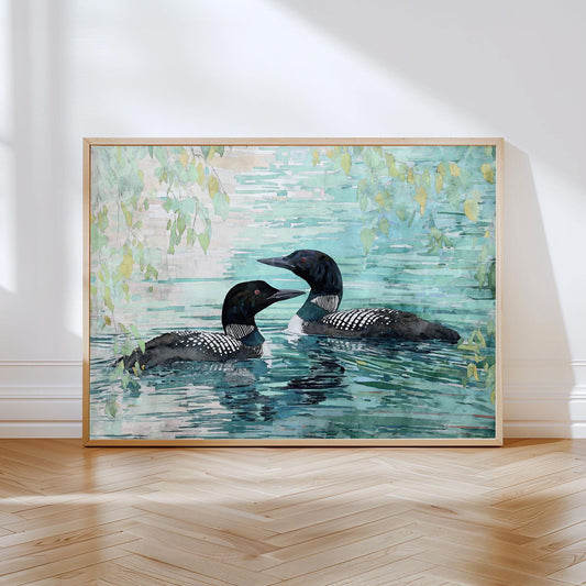 Watercolor painting of two common loons swimming side by side in calm turquoise water, framed by hanging green leaves above. The birds’ black heads, white-striped necks, and speckled wings contrast against the soft, reflective water. The artwork is displayed in a light wood frame, leaning against a white wall on a sunlit wooden floor.