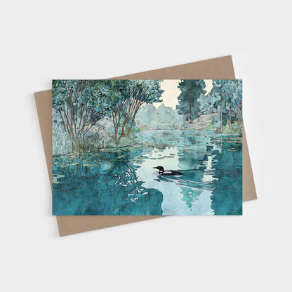 Greeting card featuring a watercolor-style illustration of a loon gliding across a calm lake. The water reflects surrounding green trees and soft blue sky, creating a serene, peaceful atmosphere. The card rests on a kraft paper envelope.