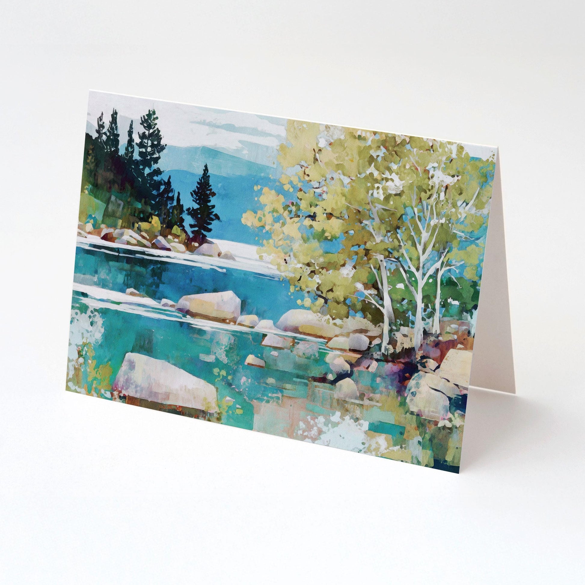 Folded greeting card standing upright, displaying a vibrant impressionist landscape of a turquoise lake with boulders, birch trees with pale trunks and green foliage on the right, and dark evergreens along the distant shoreline with soft blue mountains in the background.