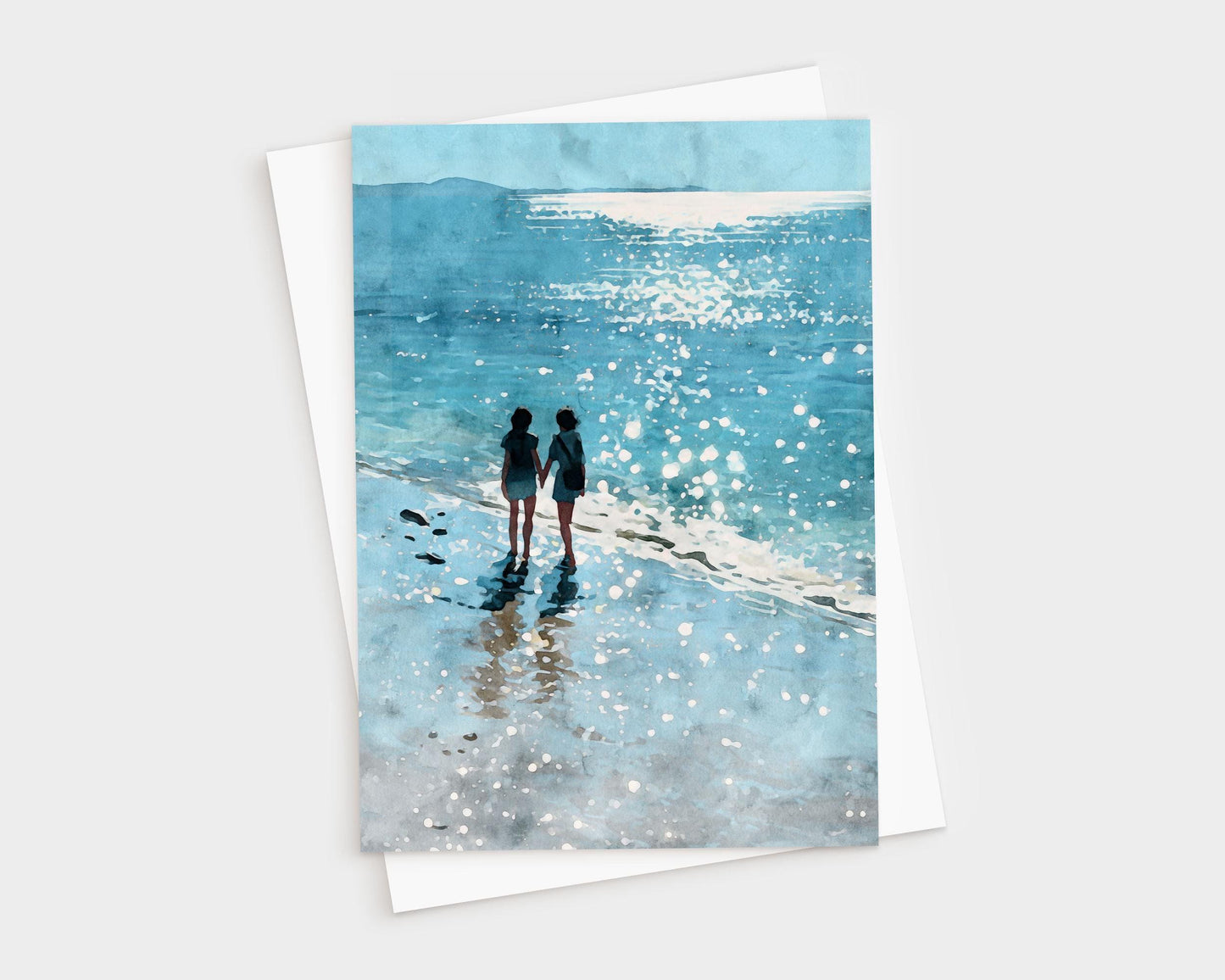 Watercolor-style greeting card featuring two people holding hands while walking along a sparkling shoreline, with their reflections in the wet sand and sunlight glittering on the ocean. A white envelope is partially visible behind the card.